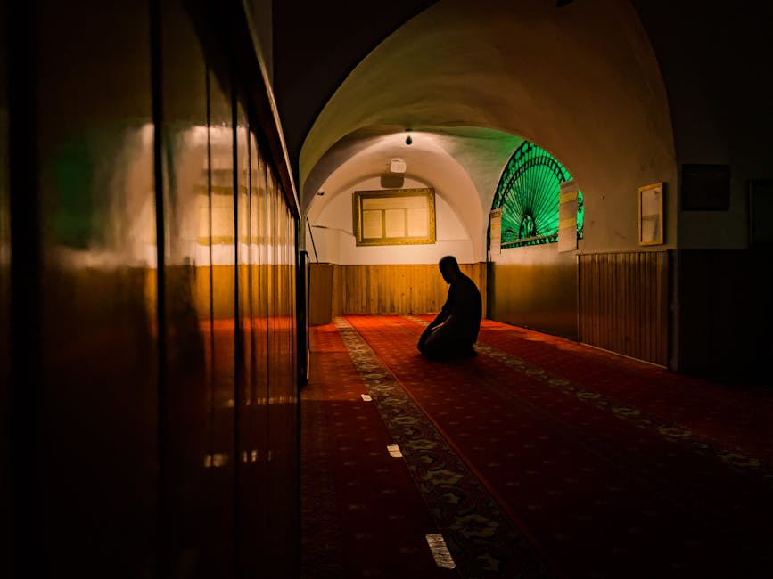 A man kneels in deep prayer inside a dimly lit mosque with arched ceilings and ornate carpet.